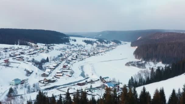 Вид з повітря на село з дерев'яними будинками на березі замерзлої річки взимку — стокове відео
