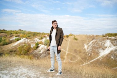 Kot pantolonlu, tişörtlü ve pelerinli ciddi bir adam tepelik arazide duruyor. Kapüşonlu gözlüklü yetişkin bir erkek kırsala dikkatlice bakıyor.