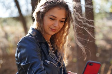 Parkta akıllı telefon kullanan genç bir kadın. Gülümseyen, uzun saçlı, rüzgârda sallanan, güz gününü parkta geçirirken cep telefonu kullanan genç bir kadın..