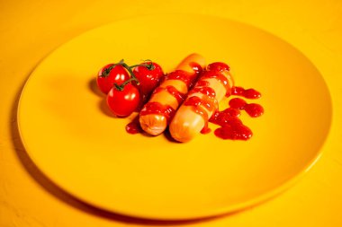 Soslu sosisler ve sarı tabakta kiraz domatesleri. Kırmızı domatesli sosis ve sarı arkaplanda ketçap.