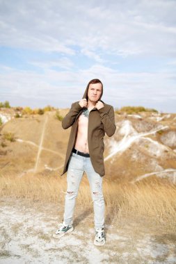 Pelerinli çıplak gövdeli dövmeli adam tepelik arazide duruyor. Yetişkin erkek bulutlu bir günde kırsalda poz verir.