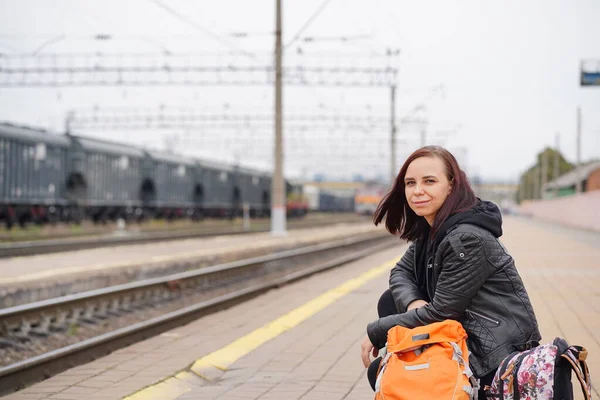 Young woman squats on platform, waiting for train. Female passenger with backpacks sitting on railroad platform in waiting for train ride