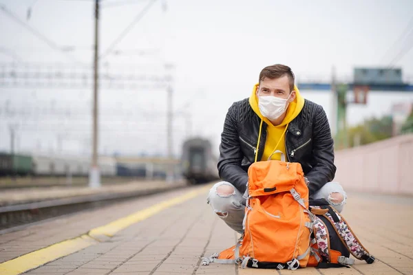 Young man in medical mask squats on platform, waiting for train. Male passenger in protective mask with backpacks sitting on railroad platform in waiting for train ride during coronavirus pandemic