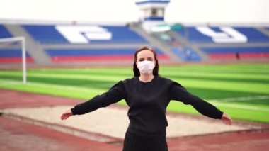 Tıbbi maskeli genç bir kadın stadyumda ısınma antrenmanı yapıyor. Siyah günlük giysiler içinde yetişkin bir kadın temiz havada spor yapıyor ve halka açık yerlerde kendini hastalıklardan koruyor.