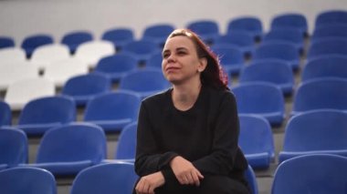 Siyah eşofmanlı, uzun saçlı genç bir kadın stadyumda tek başına oturmuş ve maç izliyor..