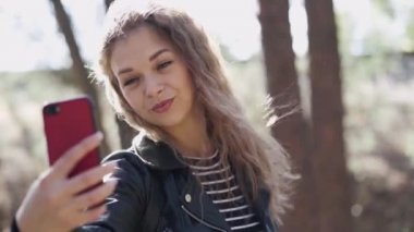 Parkta akıllı telefon kullanan genç bir kadın. Gülümseyen, uzun saçlı, rüzgârda sallanan, güz gününü parkta geçirirken cep telefonu kullanan genç bir kadın.