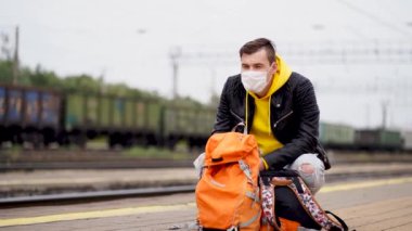 Sağlık maskeli genç adam platformda tren bekliyor. Korunaklı maskeli, sırt çantalı, koronavirüs salgını sırasında tren yolculuğunu bekleyen bir erkek yolcu..