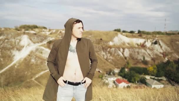 Tetovaný muž s holým trupem v plášti stojí v kopcovitém terénu. Dospělý samec má za větrného počasí krásný výhled na krajinu. Zpomalený pohyb. — Stock video