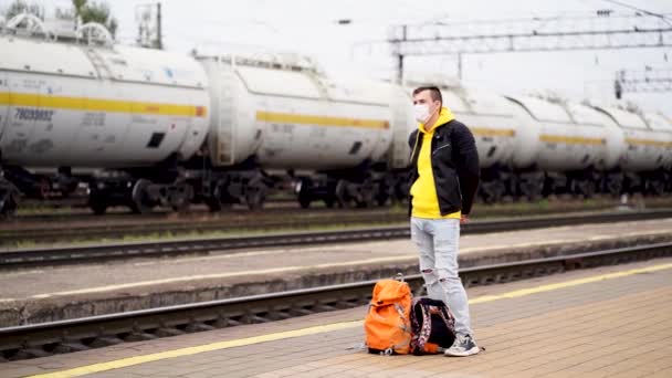 戴着医疗面具的年轻人站在站台上等火车。铁路站台上戴着护目镜、背着背包的男性乘客在验尸官大流行期间等火车. — 图库视频影像