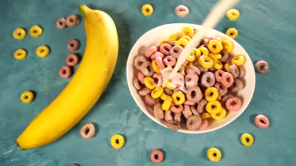 Zdrowe śniadanie z płatkami i bananem. Z góry miski z pyszne zdrowe śniadanie wykonane z kolorowych pierścieni zbożowych i banana z mlekiem serwowane na niebieskim stole — Wideo stockowe