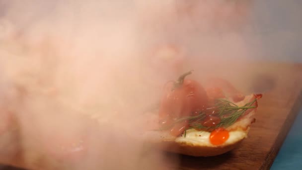 Appetitliche Bruschetta auf Holzbrett mit zerstreuendem Dampf. Nahaufnahme von Sandwich mit geschmolzenem Käse, Speck, Kirschtomaten, Dill und Sauce auf blauem Hintergrund. — Stockvideo