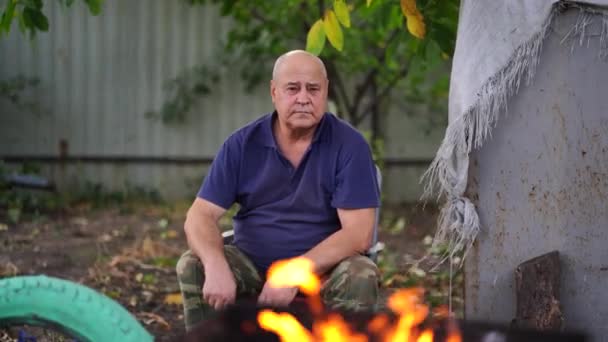 Uomo maturo seduto sulla sedia in giardino. Maschio anziano acceso fuoco in chargrill per arrostire shashlik con verdure su spiedini. Concetto di picnic all'aperto. — Video Stock