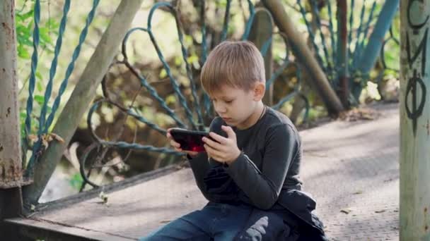 Niño enfocado usando smartphone en el parque. Niño de edad escolar concentrado serio en ropa casual sentado en el banco y jugando en el teléfono móvil mientras pasa el día de otoño en el parque — Vídeo de stock