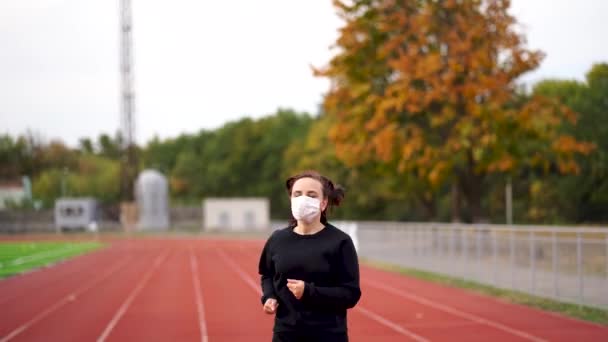 Mujer activa en máscara protectora corriendo en el estadio. Joven hembra en ropa deportiva y máscara protectora para la prevención del coronavirus corriendo en pista roja durante el entrenamiento al aire libre en el estadio — Vídeos de Stock