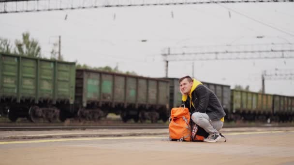 Genç adam platforma çömelmiş, treni bekliyor. Tren istasyonunda sırt çantalı bir erkek yolcu tren yolculuğu için bekliyor. Turizm, seyahat ve dinlenme kavramı. — Stok video