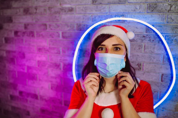 Женщина Костюме Санта Клауса Исправляет Медицинскую Маску Против Подсвеченной Стены — стоковое фото