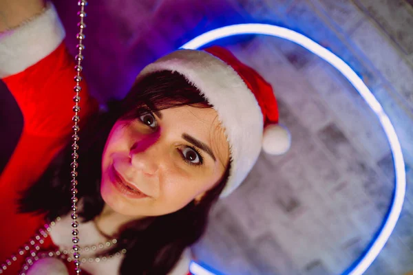 穿着圣诞老人套装 头戴珠子 靠着灯光明亮的墙壁的年轻女子的画像 戴着圣诞礼帽拿着珠子的漂亮女性的近照 圣诞庆祝的概念 — 图库照片