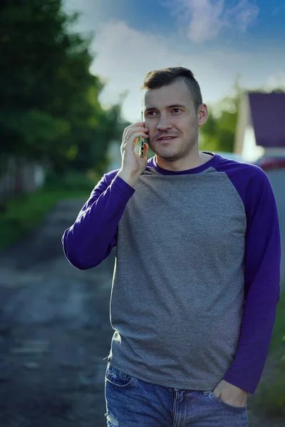 Man speaking on smartphone in suburb. Adult male in casual clothes answering phone call and looking away while standing on blurred background of suburban street.