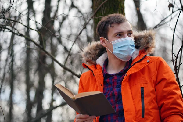 Adult man in medical mask reads book in autumn forest. Lonely male in protective mask resting with book on fresh air, enjoying his leisure time