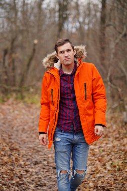 Sonbahar ormanında yürüyen turuncu ceketli genç adam. Güz mevsiminde temiz hava alan yetişkin bir erkek.