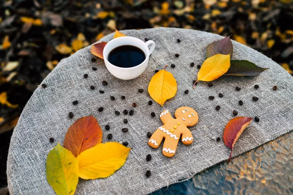 Gingerbread man and cup of coffee near autumn leaves. Top view of sweet gingerbread man and cup of strong espresso coffee placed near colorful autumn leaves on linen cloth in park