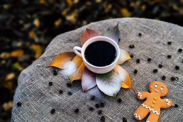 Gingerbread man and cup of coffee near autumn leaves. Top view of sweet gingerbread man and cup of strong espresso coffee placed near colorful autumn leaves on linen cloth in park.