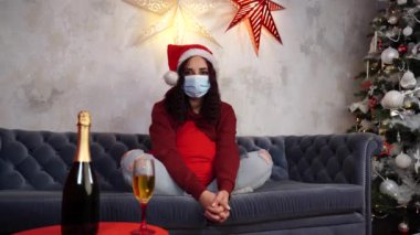 Tıbbi maskeli genç bir kadın ve Noel Baba şapkası odada kanepede oturuyor. Koronavirüs salgını sırasında güvenli bir Noel kutlaması yapan koruyucu maskeli çekici esmer kadın..