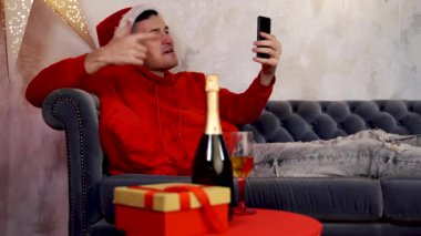 Noel Baba şapkalı genç yakışıklı bir adam bir bardak şampanyayla akıllı telefondan arkadaşlarıyla canlı yayın yapıyor. İnternette Noel 'i kutlayan ve evdeki kanepede uzanan mutlu adam..