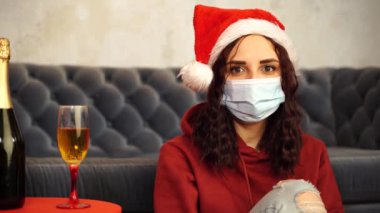 Tıbbi maskeli genç bir kadın ve odada koltuğun yanında oturan Noel Baba şapkası. Koronavirüs salgını sırasında güvenli bir Noel kutlaması yapan koruyucu maskeli çekici esmer kadın..