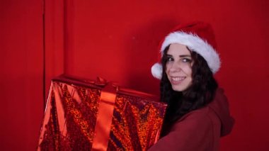 Noel Baba şapkalı genç kadın kırmızı duvarın yanında duran büyük bir hediye tutuyor. Noel şapkalı, hediye kutulu mutlu bir kadın. Tatil kavramı, hediyeler ve iyi ruh hali..