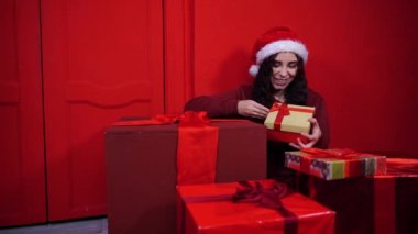 Noel Baba şapkalı genç bir kadın kırmızı duvarın yanında hediyeler içinde oturuyor. Çekici esmer tebessüm, kameraya bakıyor. Tatil kavramı, hediyeler ve iyi ruh hali..