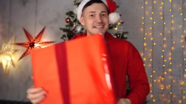 Noel Baba şapkalı genç yakışıklı adam Noel ağacının arkasında büyük bir hediyeyle dans ediyor. Mutlu adam dans ediyor, neşeli hediyeler veriyor. Tatil kavramı, hediyeler ve iyi ruh hali..