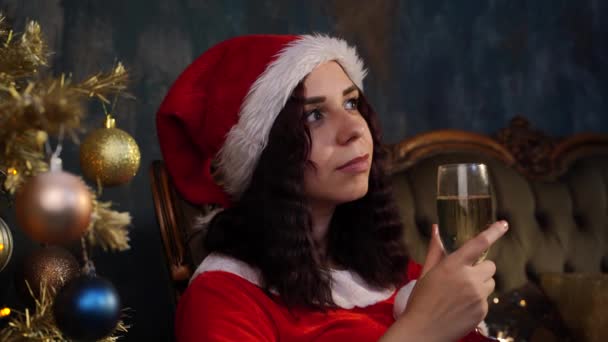サンタクロースの衣装を着た若い思慮深い女性で クリスマスツリーの近くの椅子にシャンパンのガラスが座っています 自宅でクリスマスを祝う夢のような女性 休日と良い気分の概念 — ストック動画