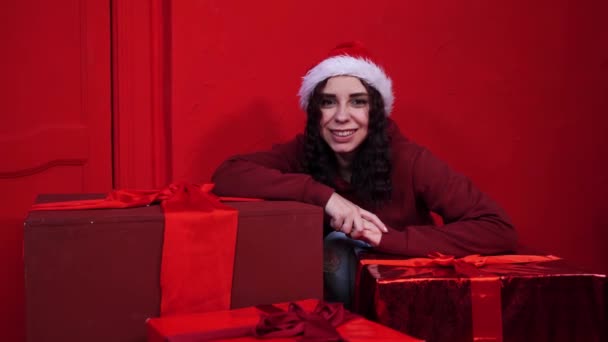 サンタクロースの帽子の若い女性は赤い壁の近くの贈り物に座っている 魅力的なブルネットの笑顔 カメラを見て 贈り物と良い気分の概念 — ストック動画