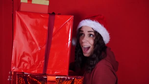 サンタクロースの帽子の若い女性は 赤い壁の近くに立って 大きな贈り物を保持しています ギフトボックス付きクリスマスの帽子の幸せな女性 プレゼントと良い気分の概念 — ストック動画