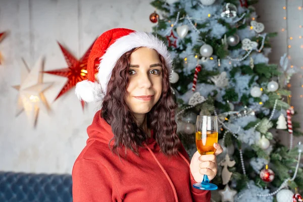 年轻的女人戴着圣诞老人的帽子 喝着香槟 站在圣诞树旁 迷人的黑发姑娘摆设在针叶树旁 装饰华丽 在家里庆祝圣诞节的概念 — 图库照片