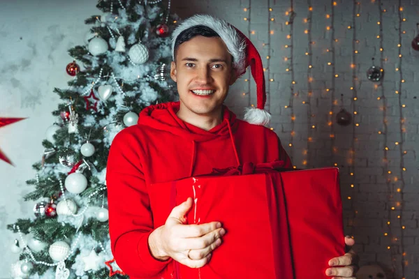 サンタクロースの帽子の若いハンサムな男は クリスマスツリーを背景に大きな贈り物で踊ります 幸せな男が踊り 現在を楽しんでいます プレゼントと良い気分の概念 — ストック写真