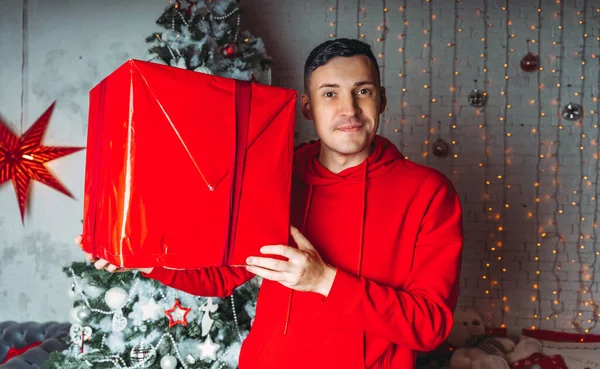 クリスマスツリーを背景に大きなラップギフトを持つ若いハンサムな男 かなりの男が箱を持ってる プレゼントと良い気分の概念 — ストック写真