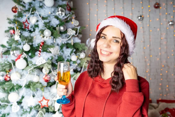 年轻的女人戴着圣诞老人的帽子 喝着香槟 站在圣诞树旁 迷人的黑发姑娘摆设在针叶树旁 装饰华丽 在家里庆祝圣诞节的概念 — 图库照片