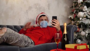 Tıbbi maskeli ve Noel Baba şapkalı bir adam akıllı telefondan arkadaşlarıyla canlı yayın yapıyor. Odadaki kanepede yatıyor. Coronavirus salgını sırasında internette şampanyalı adam Noel 'i kutluyor..