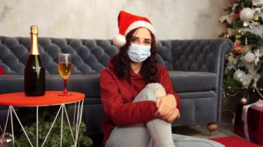 Tıbbi maskeli genç bir kadın ve odada koltuğun yanında oturan Noel Baba şapkası. Koronavirüs salgını sırasında güvenli bir Noel kutlaması yapan koruyucu maskeli çekici esmer kadın..
