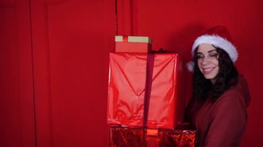 Noel Baba şapkalı genç kadın kırmızı duvarın yanında duran büyük hediyeler taşıyor. Noel şapkalı, hediye kutularıyla mutlu bir kadın. Tatil kavramı, hediyeler ve iyi ruh hali..