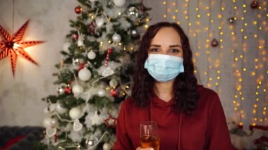 Tıbbi maskeli genç bir kadın ve Noel Baba şapkası ve arka planda bir bardak şampanya. Koronavirüs salgını sırasında güvenli Noel 'i kutlayan koruyucu maskeli çekici esmer..