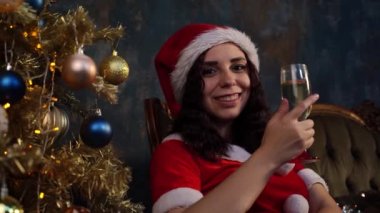 Noel Baba kostümlü genç bir kadın, elinde bir bardak şampanya, Noel ağacının yanındaki sandalyede oturuyor. Çekici kadın gülümsüyor, kameraya bakıyor. Evde Noel kutlaması kavramı.