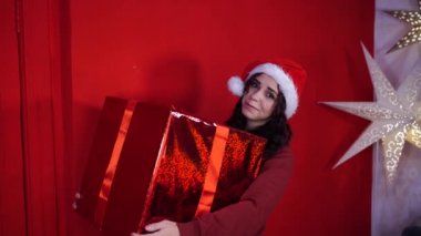 Noel Baba şapkalı genç kadın kırmızı duvarın yanında duran büyük bir hediye tutuyor. Noel şapkalı, hediye kutulu mutlu bir kadın. Tatil kavramı, hediyeler ve iyi ruh hali..