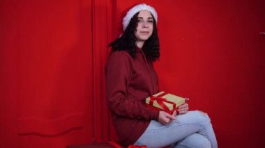 Noel Baba şapkalı çekici bir kadın kırmızı duvarın yanında hediyelerin üzerinde oturuyor. Gülümseyen kadın hediye kutusunun üzerinde oturuyor. Tatil kavramı, hediyeler ve iyi ruh hali..