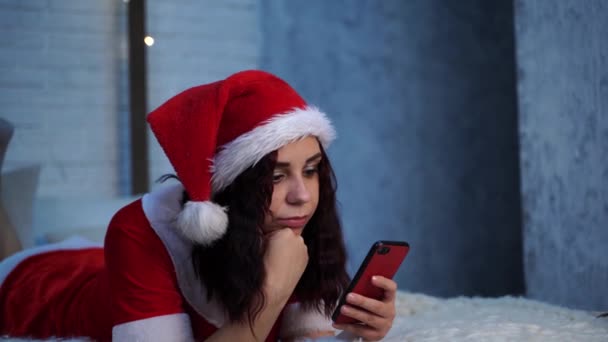 身穿圣诞老人服装的年轻女子躺在床上浏览智能手机 迷人的女性休息与手机和庆祝圣诞节在家里 假日的概念和良好的心情 — 图库视频影像