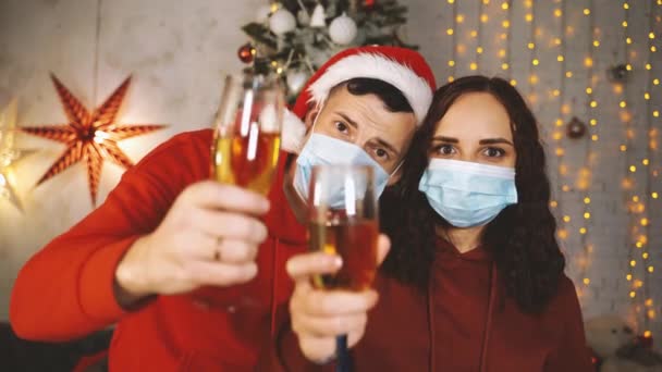 医療用マスクの若い男性と女性は クリスマスツリーを背景にシャンパンのグラスを叩いて トーストを提供しています コロナウイルスのパンデミックの間の安全なクリスマスを祝う夫婦 — ストック動画