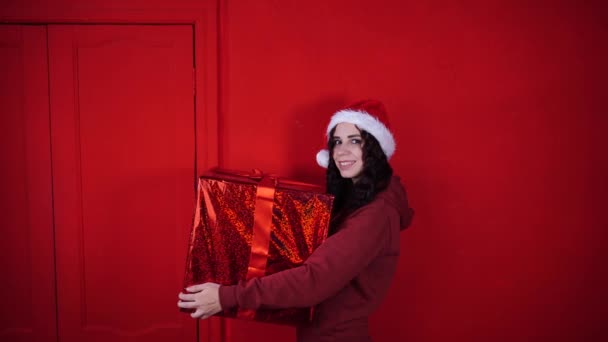 戴着圣诞老人帽子的年轻女子站在红墙边 手里拿着一份大礼物 戴着圣诞礼帽 头戴圣诞礼帽的快乐女性 送礼和心情好的概念 — 图库视频影像