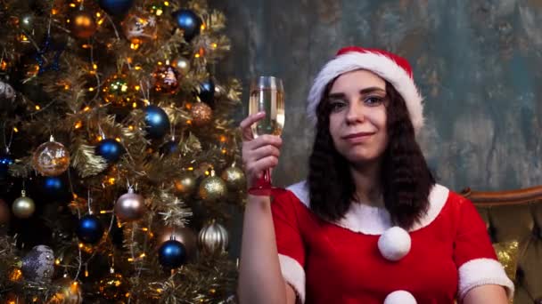 身穿圣诞老人服装 拿着香槟的年轻女子坐在圣诞树旁的椅子上 迷人的女性微笑着 看着相机 在家里庆祝圣诞节的概念 — 图库视频影像
